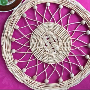 May include: A round woven wicker coaster with a decorative design of woven circles and wooden beads around the edge. The coaster is on a pink surface.