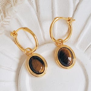 Puede incluir: Un par de pendientes de aro dorados con colgantes ovalados de piedra ojo de tigre marrón. Los pendientes se exhiben sobre una superficie blanca con forma de flor, mostrando el diseño y el color de la joyería.