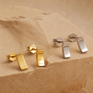 May include: Two pairs of stud earrings. One pair is gold-colored, and the other is silver-colored. Each earring has a rectangular shape with a slight curve. The earrings are displayed on a light brown, textured surface.