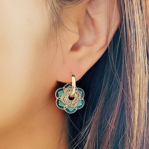 May include: Gold hoop earrings with a green and gold flower charm. The flower charm has a detailed design and is a beautiful accent to the earrings.