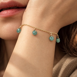 Op de afbeelding: Een goudkleurige kettingarmband met vier kleine, ronde, lichtgroene stenen bedels die aan de schakels hangen. Elke steen is vastgezet met een gouden draadwikkeling, wat een delicaat bungelend effect langs de ketting geeft.