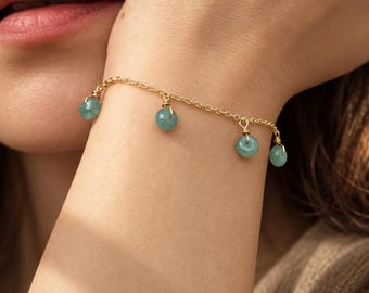 May include: A gold chain bracelet featuring four small, round, light green stone charms suspended from the links. Each stone is secured with a gold wire wrap, creating a delicate dangling effect along the wrist chain.