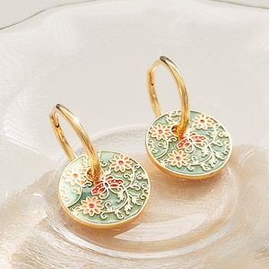 May include: A pair of gold hoop earrings with a green and white enamel floral design. The earrings are round and decorated with small red flowers.