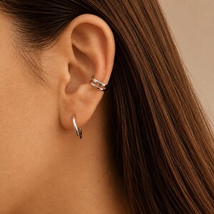 May include: A close-up of an ear featuring silver jewellery. A small hoop earring is in the earlobe, and a double-banded ear cuff is on the upper ear. The jewellery is shiny and reflective, set against the backdrop of brown hair.
