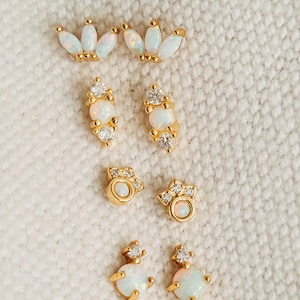 Könnte beinhalten: Eine Sammlung goldfarbener Ohrringe mit weißen Opal- und klaren Kristallakzenten. Die Ohrringe weisen verschiedene Designs auf, darunter Kronenformen, vertikale Cluster und runde Ohrstecker, vor einem strukturierten weißen Hintergrund.