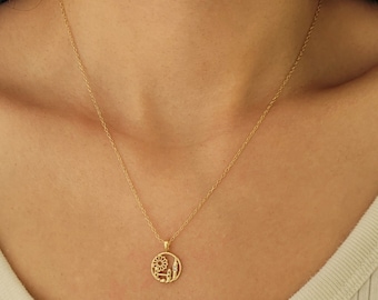 Puede incluir: Un collar dorado con un colgante circular. El colgante presenta un sol, edificios y otros elementos arquitectónicos. El collar se lleva alrededor del cuello.