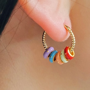 Puede incluir: Aretes de aro de oro con cuentas de esmalte de colores. Las cuentas son de color morado, azul, naranja claro, verde y rojo. Cada cuenta tiene pequeños puntos dorados.