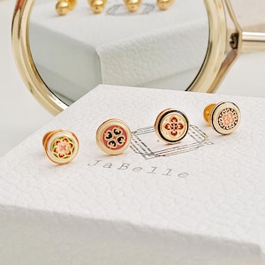 May include: Four gold-toned cufflinks with colourful enamel designs. Each cufflink features a different geometric pattern with red, green, black, and white accents. The cufflinks are displayed on a white box with the text "JaBelle" printed on the front.