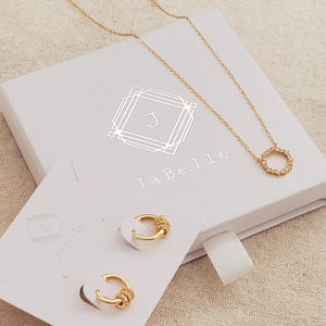 May include: A white gift box with gold lettering that says "JaBelle" and a gold initial "J" in a decorative frame. The box contains a gold necklace with a small circle pendant and a pair of gold hoop earrings. The box is on a beige fabric surface.