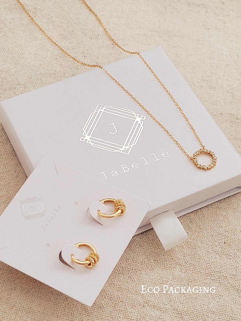 May include: A white gift box with gold lettering that says "JaBelle" and a gold initial "J" in a decorative frame. The box contains a gold necklace with a small circle pendant and a pair of gold hoop earrings. The box is on a beige fabric surface.