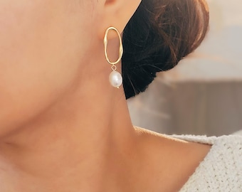 May include: Gold earrings with a dangling pearl. The earrings are shaped like an oval with an irregular, slightly wavy edge.
