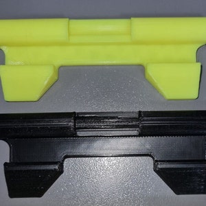 Może przedstawiać: Dwa obiekty drukowane w 3D. Jeden jest jasnożółty, drugi czarny. Oba mają kształt prostokątnego pręta z podniesioną częścią środkową i skośnymi, trapezowymi końcami. Są to elementy lub części większego zespołu.