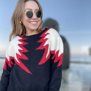 Puede incluir: Suéter de punto negro y blanco con un patrón de rayos rojos. El suéter tiene un cuello redondo y mangas largas.