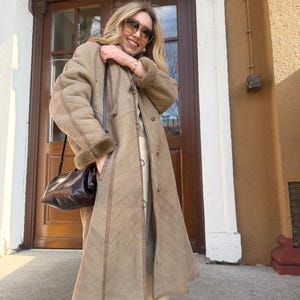 May include: A long, tan coat with a shearling lining and a diagonal stitched pattern. The coat has a button-up front, a hood, and a dark brown leather handbag. The coat is worn outdoors in front of a building.
