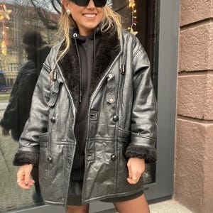 May include: A black leather jacket with a fur collar and cuffs. The jacket has a button closure and a belt at the waist. The model is wearing a black hoodie and a brown baseball cap with the New York Yankees logo.