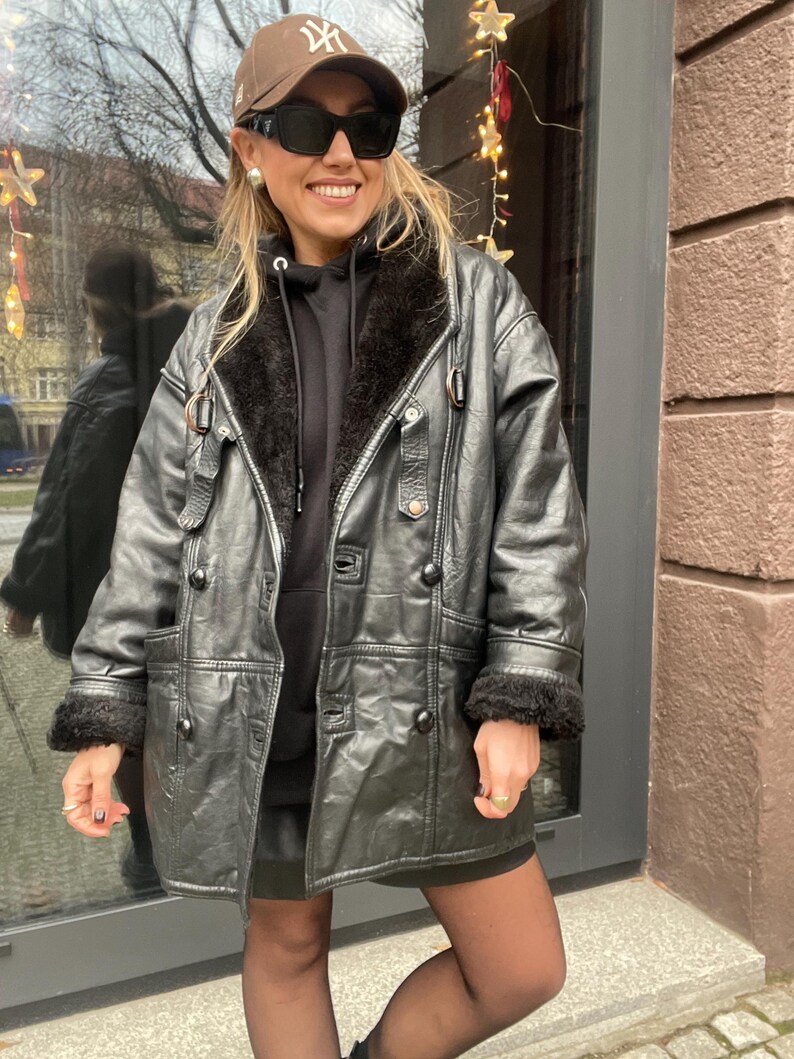 May include: A black leather jacket with a fur collar and cuffs. The jacket has a button closure and a belt at the waist. The model is wearing a black hoodie and a brown baseball cap with the New York Yankees logo.