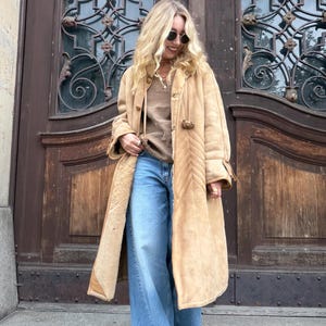 Könnte beinhalten: Ein langer, beigefarbener Wildledermantel mit weichem Innenfutter, Knebelverschlüssen und Chevron-Steppnähten. Der Mantel wird über einem braunen Oberteil und einer weiten blauen Jeans getragen. Das Kleidungsstück hat lange Ärmel und einen Kragen.