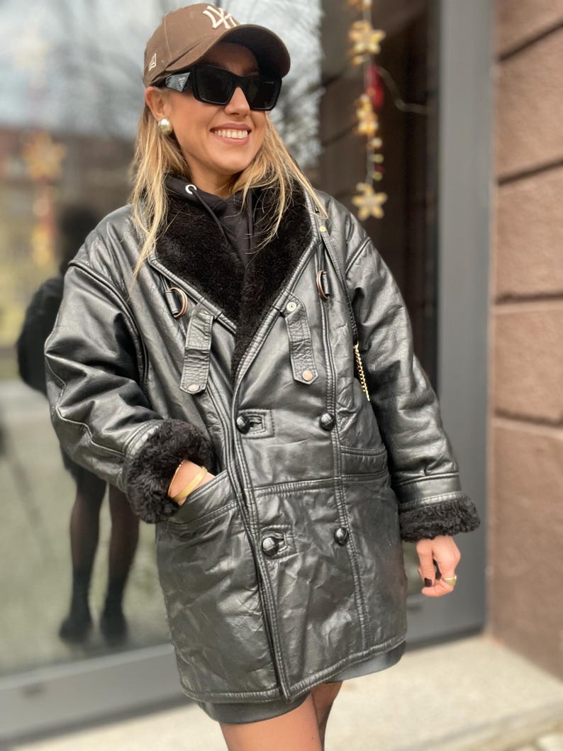 May include: A black leather jacket with a fur collar and a double-breasted closure. The jacket has a gold buckle on the belt and a gold button on the pocket.