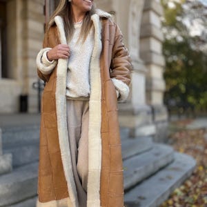 Può includere: Un lungo cappotto in montone marrone con fodera e finiture in montone color crema. Il cappotto presenta un design classico con colletto a revers e tasche applicate. La persona indossa occhiali da sole e un maglione.