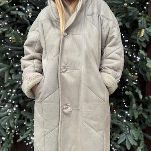 May include: A light beige, long coat with a shearling lining and a button-up front. The coat has a quilted design and a large collar. The cuffs are turned up to reveal the shearling lining. The person is wearing sunglasses and a beanie.