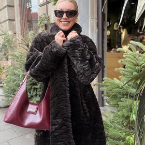 Könnte beinhalten: Eine Frau trägt einen dunkelbraunen Kunstfellmantel und eine schwarze Sonnenbrille und hält eine bordeauxfarbene Handtasche. Der Mantel hat ein strukturiertes Muster. Die Handtasche hat eine rechteckige Form und einen Schulterriemen. Der Hintergrund umfasst Grün und ein Gebäude.