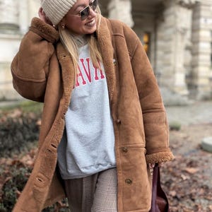 Puede incluir: Un abrigo de borrego marrón, un gorro crema y gafas de sol. El abrigo tiene una botonadura y un forro de piel. Se ve una sudadera gris con la palabra "AVA" en rojo. El conjunto se completa con pantalones de cuadros y un bolso tote burdeos.
