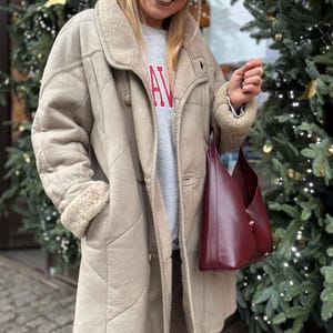 May include: A beige shearling coat with a cream shearling collar and cuffs. The coat features a double-breasted design and a quilted pattern. A burgundy leather tote bag is held. The person wears a grey sweatshirt with the letters "AV" in red.