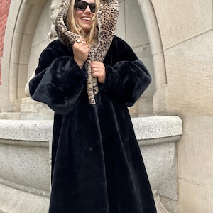 May include: A long, black faux fur coat with a hood lined with a leopard print pattern. The coat has large sleeves and a button closure. The person in the image is wearing sunglasses and holding the hood up.