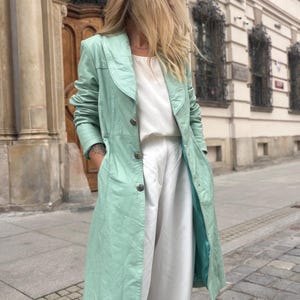 May include: A long, mint green leather coat featuring a wide collar, long sleeves, and a front button closure. The coat is worn over a plain white top and white trousers, creating a layered look with a smooth, structured finish and visible seams.