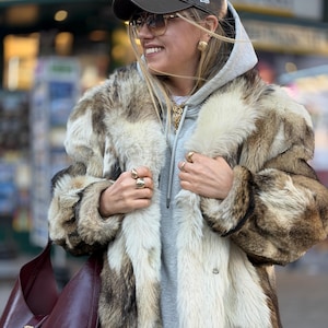 May include: A faux fur coat in shades of brown, cream, and gray. The coat has a wide collar and long sleeves. The person is wearing a gray hoodie, a brown baseball cap, and sunglasses. A burgundy leather shoulder bag is visible.