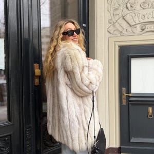 Puede incluir: Una mujer con un abrigo de piel claro, gafas de sol negras y un bolso negro. El abrigo tiene un aspecto texturizado. La mujer está de pie frente a una puerta oscura.