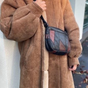 Könnte beinhalten: Ein beigefarbener Wildledermantel mit cremefarbenem Lammfellfutter und eine kleine, halbmondförmige Umhängetasche aus einem Patchwork-Design aus schwarzen und dunkelbraunen Lederteilen mit einem dünnen schwarzen Riemen und sichtbaren Nähten.