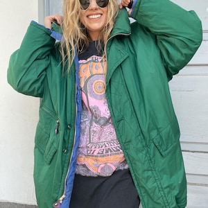 Può includere: Una giacca trapuntata verde con fodera blu e collo alto. La giacca ha una chiusura a zip e tasche. La persona indossa una maglietta grafica e occhiali da sole.