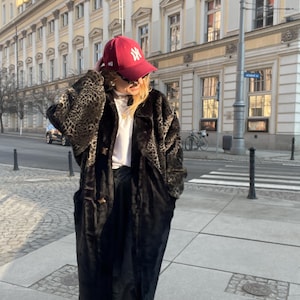 Könnte beinhalten: Eine Frau trägt einen schwarzen und braunen Leopardenfell-Mantel aus Kunstfell mit einer roten Baseballkappe, auf der "NY" steht. Sie trägt ein weißes T-Shirt und eine schwarze Hose.