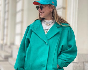 May include: A teal blue wool jacket with a double breasted closure and large pockets. The jacket has a relaxed fit and is worn with a white t-shirt and a teal blue baseball cap with a white logo.