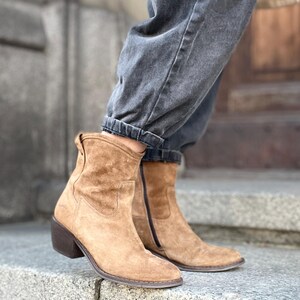 Pu&ograve; includere: Stivaletti in camoscio color cammello con tacco a blocco. Gli stivali hanno una cerniera laterale e una punta arrotondata. Gli stivali sono indossati con jeans di denim grigio scuro. Gli stivali sono alti circa 15 cm.