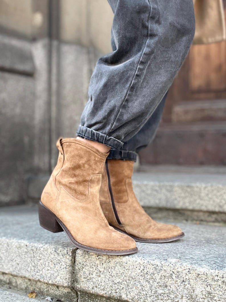 Pu&ograve; includere: Stivaletti in camoscio color cammello con tacco a blocco. Gli stivali hanno una cerniera laterale e una punta arrotondata. Gli stivali sono indossati con jeans di denim grigio scuro. Gli stivali sono alti circa 15 cm.