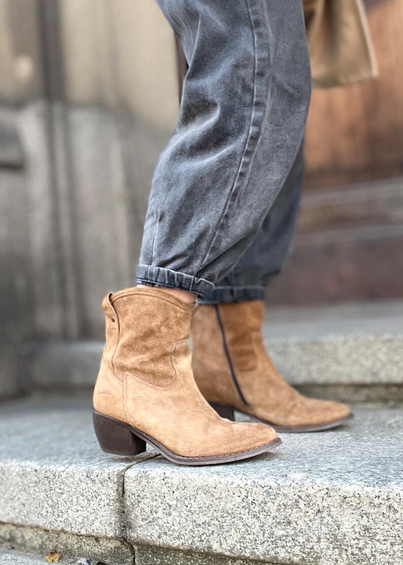 Pu&ograve; includere: Stivaletti in camoscio color cammello con punta affilata e tacco a blocco. Gli stivali hanno un design di ispirazione western e sono indossati con jeans di denim grigio scuro. Gli stivali sembrano essere realizzati in un materiale morbido e strutturato.