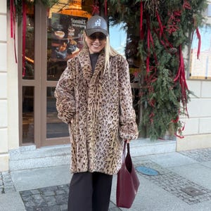 Peut inclure: Une femme porte un manteau à imprimé léopard, un pantalon noir et une casquette de baseball noire avec un logo "A". Elle tient un sac fourre-tout bordeaux et porte des lunettes de soleil. En arrière-plan, une devanture de magasin décorée pour les fêtes.