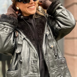 May include: A black leather jacket with a fur-lined hood. The jacket has a zipper closure and multiple pockets. The jacket is worn by a person wearing a black hoodie and a brown baseball cap.