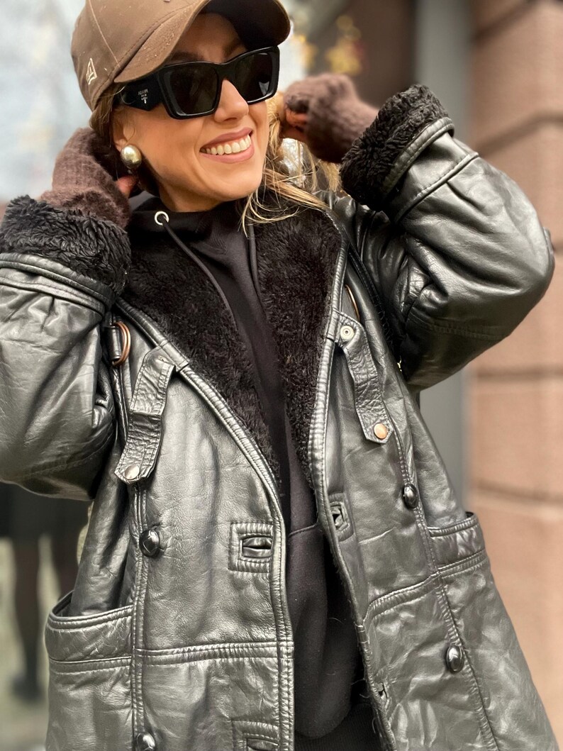 May include: A black leather jacket with a fur-lined hood. The jacket has a zipper closure and multiple pockets. The jacket is worn by a person wearing a black hoodie and a brown baseball cap.