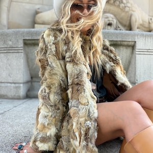May include: A long-sleeved coat made of multi-toned brown and tan fur patches, featuring a textured, shaggy appearance. The coat is worn with a white cowboy hat, dark-tinted sunglasses, a dark top, and tan leather boots.