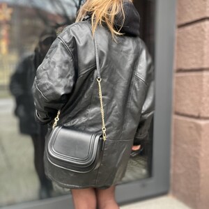 May include: A black leather jacket with a gold chain strap and a black leather handbag. The jacket has a hood and is worn over a black shirt.