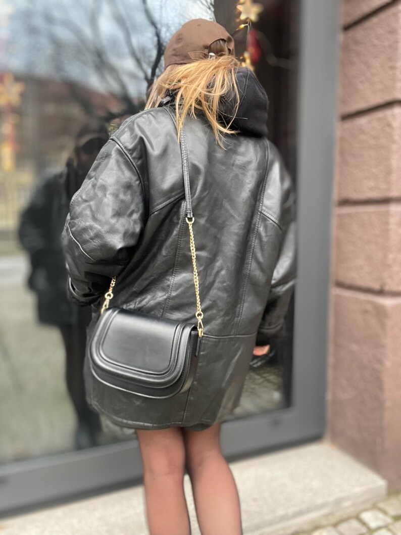 May include: A black leather jacket with a gold chain strap and a black leather handbag. The jacket has a hood and is worn over a black shirt.