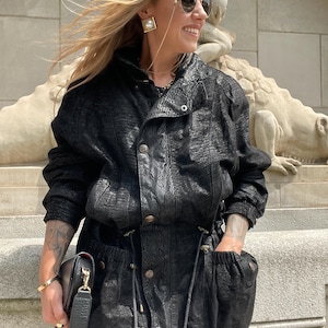 Peut inclure: Veste noire et pantalon assorti avec un motif texturé. La veste a une fermeture boutonnée sur le devant et une taille à cordon. Un sac à main noir avec une bandoulière dorée est tenu dans la main gauche. La personne porte des lunettes de soleil et des bijoux dorés.