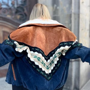 May include: A blue suede jacket with a brown suede yoke and a cream-colored shearling collar. The back features a patterned knit design in cream, green, and black. The jacket has a black ribbed hem and cuffs.