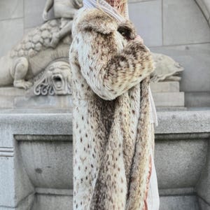 May include: A long, faux fur coat in shades of beige and brown with a spotted pattern. The coat has a plush texture and a long, flowing design. The person is wearing a baseball cap and a hooded scarf.