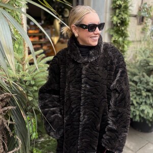 May include: A woman wearing a long, dark brown faux fur coat with a textured pattern. She is also wearing black sunglasses and a black turtleneck. The coat has a high collar and a button closure. The woman is smiling.