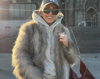 May include: A person wearing a gray hoodie, sunglasses, and a baseball cap with the letter "A" on it. They are also wearing a long, faux fur coat and carrying a burgundy handbag. The background features a large stone building.