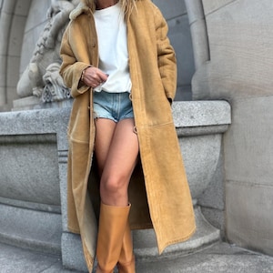 Puede incluir: Un abrigo largo de ante color tostado con cuello de piel sintética color crema, usado sobre una camiseta blanca lisa y pantalones cortos de mezclilla azul claro. El conjunto se completa con botas de cuero tostado hasta la rodilla y gafas oscuras.